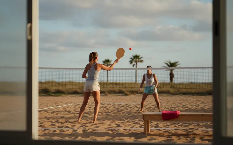 Duas jogadoras de beach tennis numa quadra de areia ao fim da tarde, vistas pela porta de vidro, com palmeiras atrás da rede
